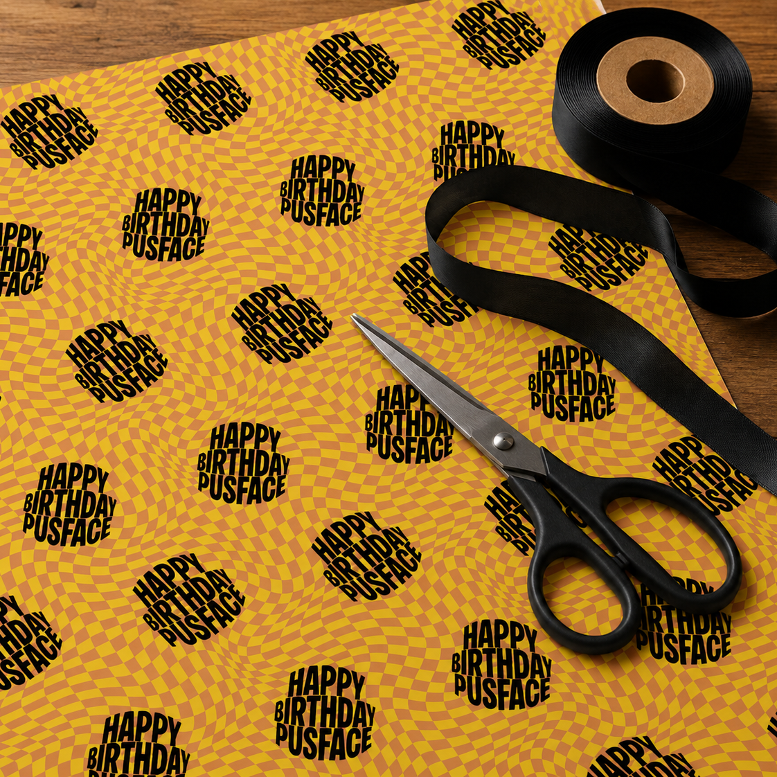 Flat lay of Happy Birthday Pusface wrapping paper on a wooden table with black ribbon and scissors ready for gift wrapping.