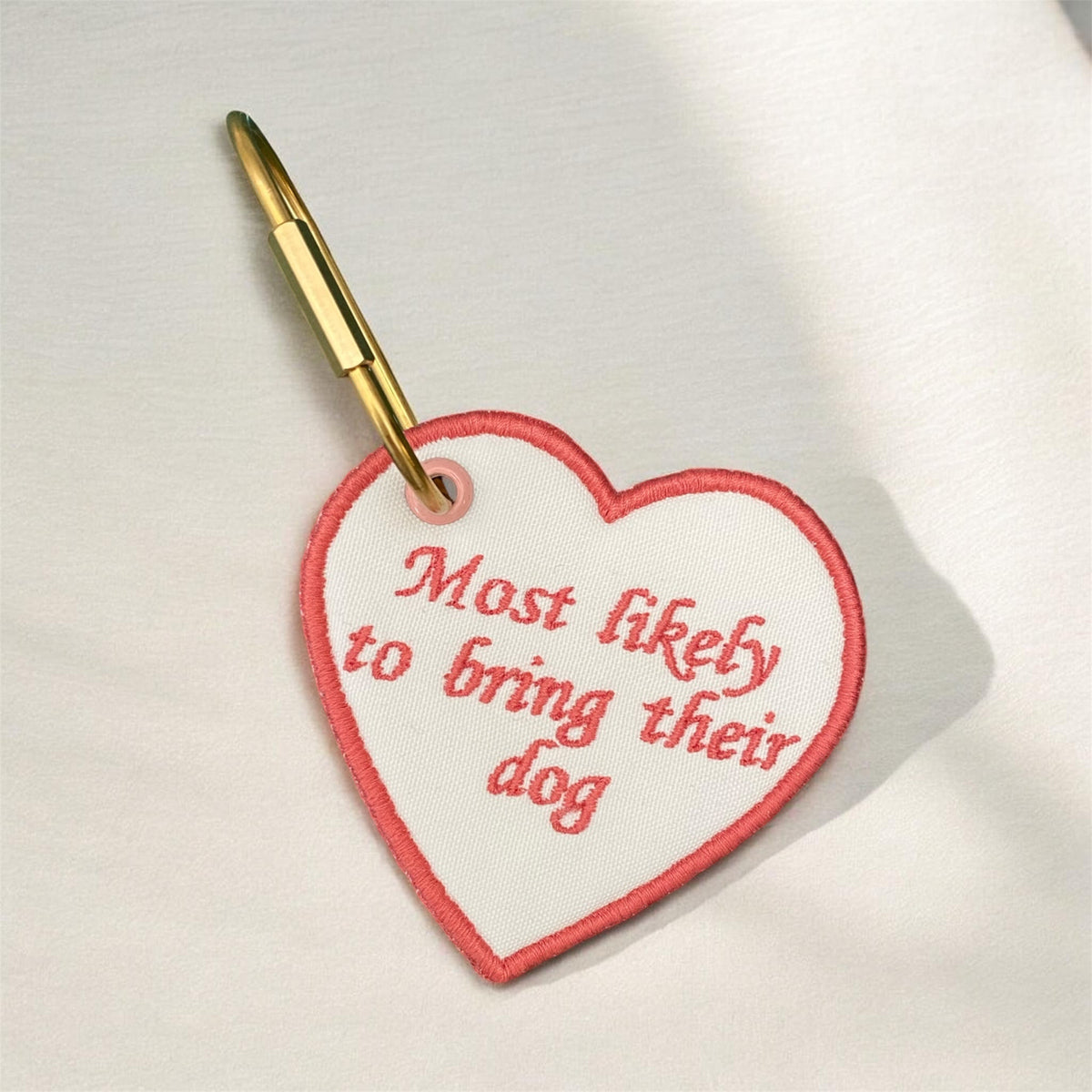 A heart-shaped embroidered patch with a gold metal clip, featuring the text 'Most likely to bring their dog' in red embroidery on a white background.