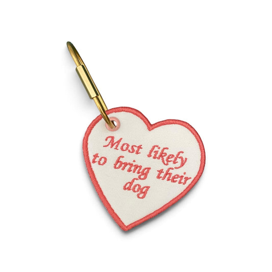 A heart-shaped white pet tag with a red stitched border and red embroidered text reading 'Most likely to bring their dog', attached to a gold metal clip, shown on a plain white background.
