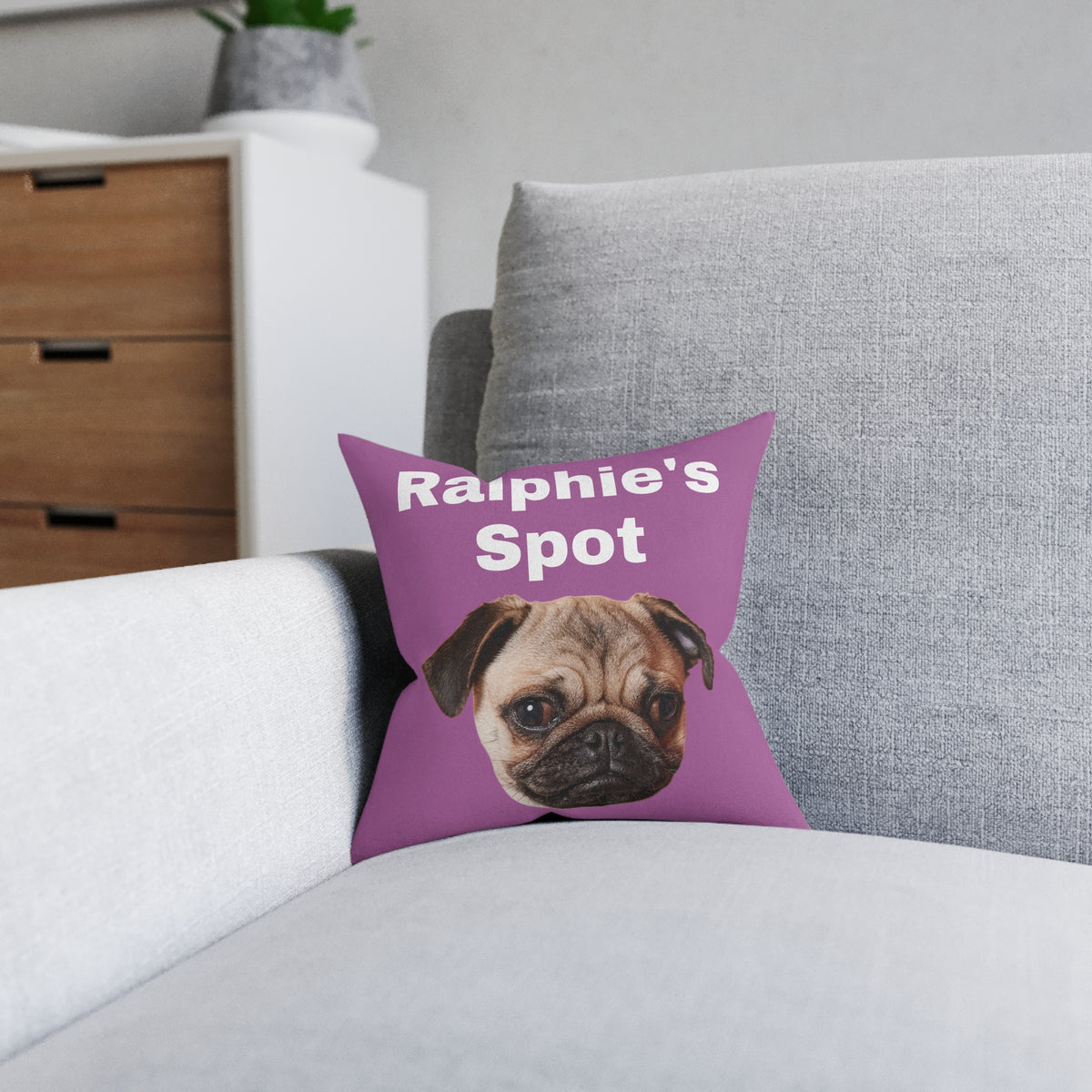 A square purple throw pillow with the text 'Ralphie's Spot' and a printed pug dog face, resting on a light gray sofa in a living room setting.