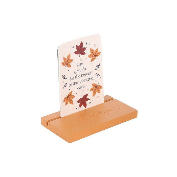 A small wooden tabletop card holder with a light natural wood base and a single upright card featuring a cream background, black script text, and scattered autumn leaf illustrations in orange, red, and brown.
