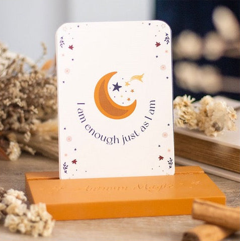 A vertical white card with a crescent moon and star illustration and the phrase 'You are enough just as you are' displayed on a wooden stand, surrounded by dried flowers and cinnamon sticks.