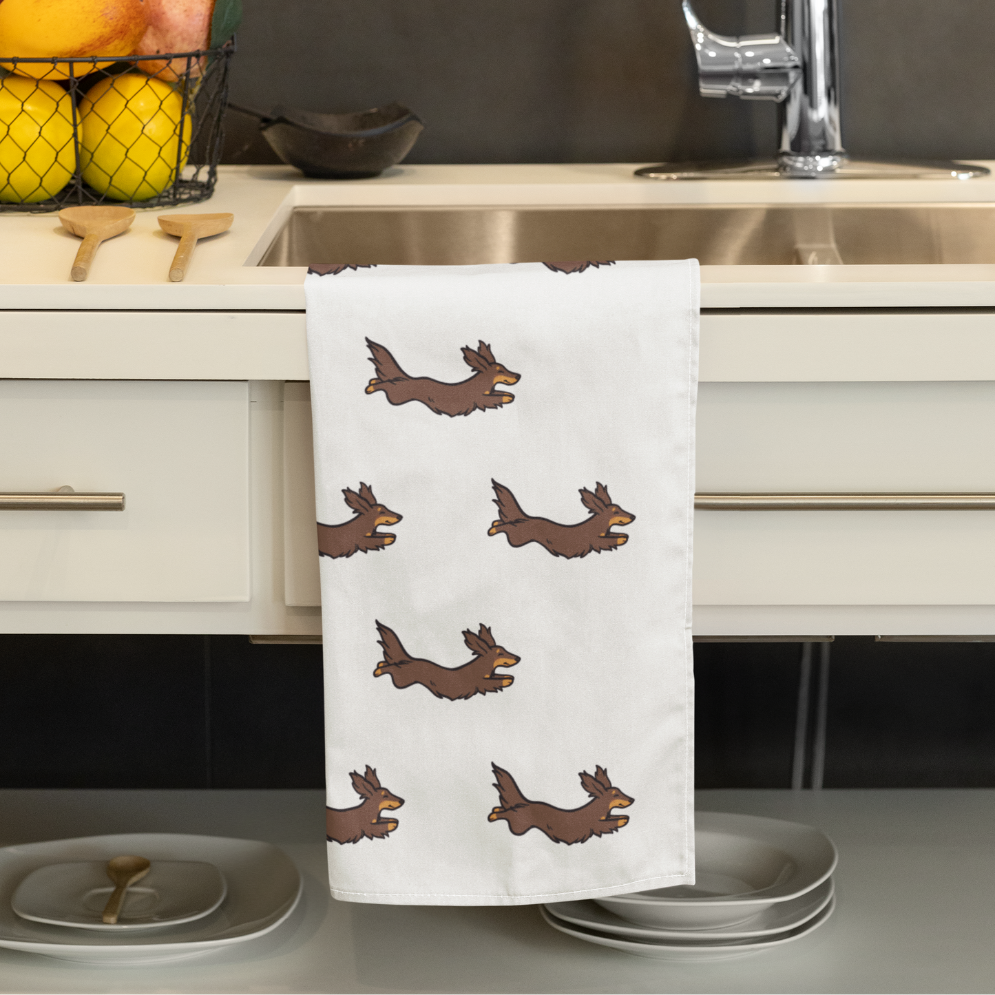 A white kitchen towel with a repeating brown dachshund pattern hanging over a stainless steel sink in a modern kitchen setting.