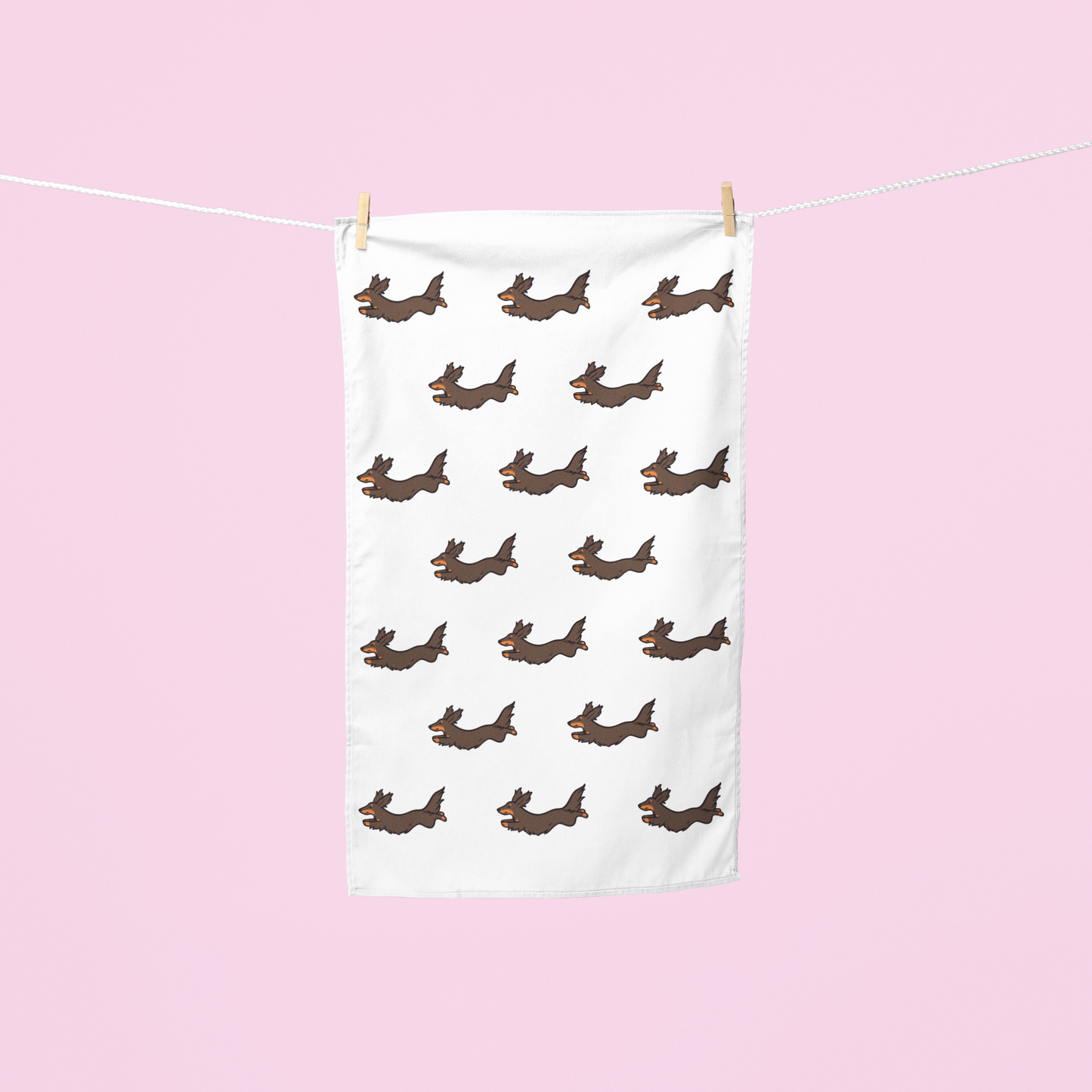 A white tea towel with a repeating pattern of brown dachshund dogs hanging from a white clothesline with wooden clothespins against a solid light pink background.