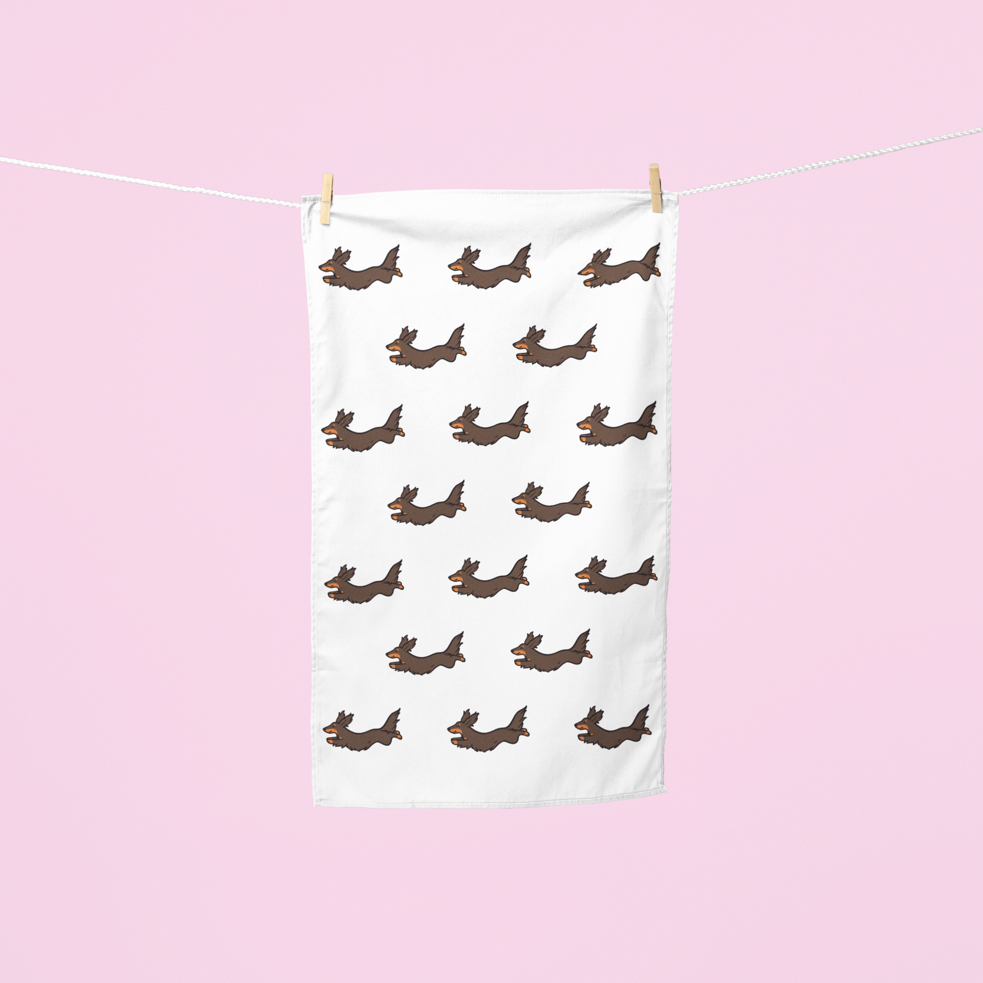 A white tea towel with a repeating pattern of brown dachshund dogs hanging from a white clothesline with wooden clothespins against a solid light pink background.