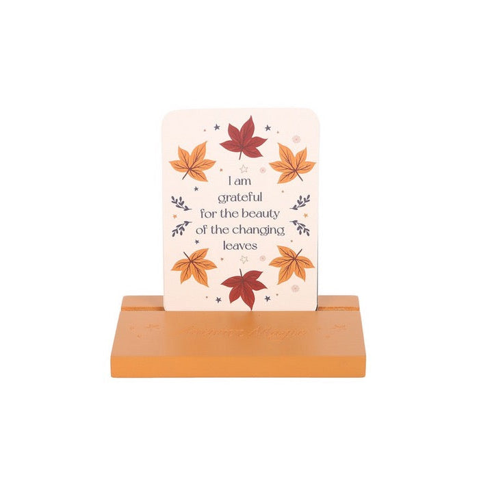 A square art print with rounded corners displayed on a small wooden stand against a plain white background. The print features a light cream background decorated with autumn maple leaves in orange, red, and yellow, alongside the quote 'I am grateful for the beauty of the changing leaves'.